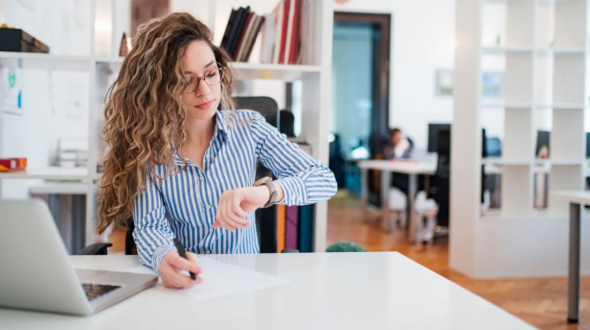 Młoda kobieta w okularach i koszuli w paski patrzy na zegarek, siedzi przy biurku w nowoczesnym biurze z laptopem i dokumentami