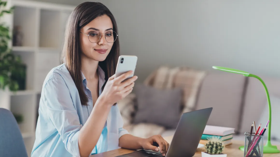 Kobieta w okularach sprawdza telefon przy laptopie